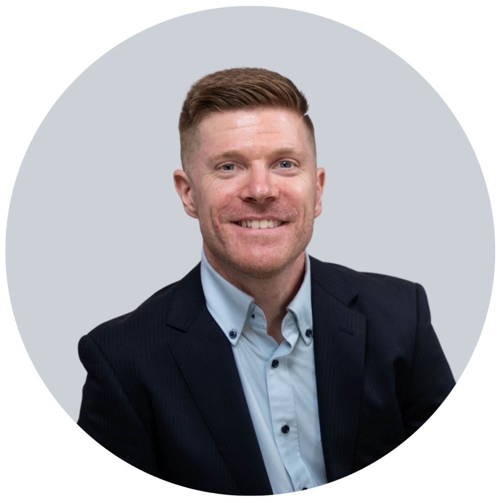 A man with short light brown hair, wearing a dark suit jacket and light blue collared shirt, smiles at the camera against a light gray background—perfect for a professional Single Page profile or Elementor template.