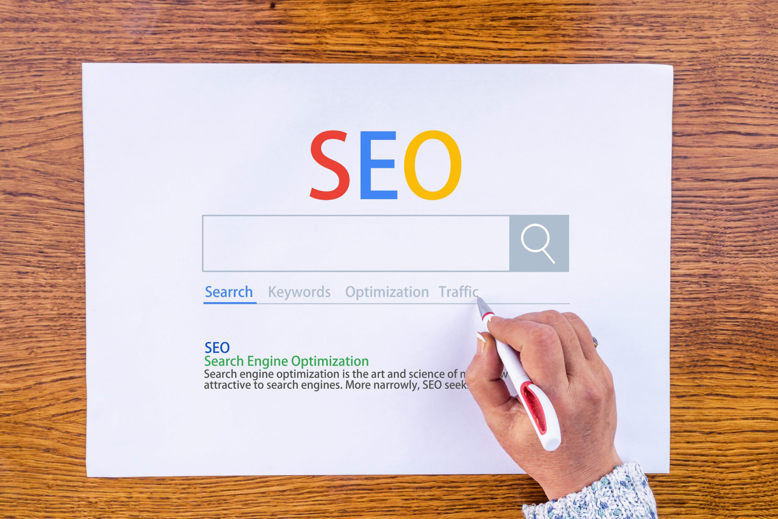 A hand holds a pen near a paper featuring the word "SEO in Orlando" above a mock search bar and search-related terms on a wooden table.