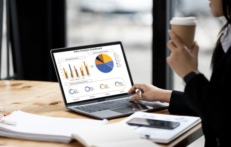 Person holding a coffee cup and using a laptop displaying a sales analysis dashboard with bar, pie, and line charts for website SEO performance during the summer season. Papers, a pen, and a phone are on the wooden table.