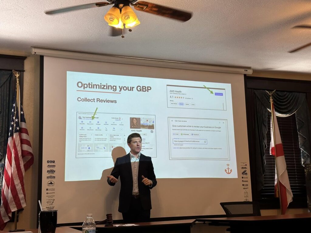 A man in a suit presents in front of a projector screen showing slides about optimizing Google Business Profile by collecting reviews, highlighting strategies used by Anchor Online Web Design. Two flags and a ceiling fan are visible in the room.