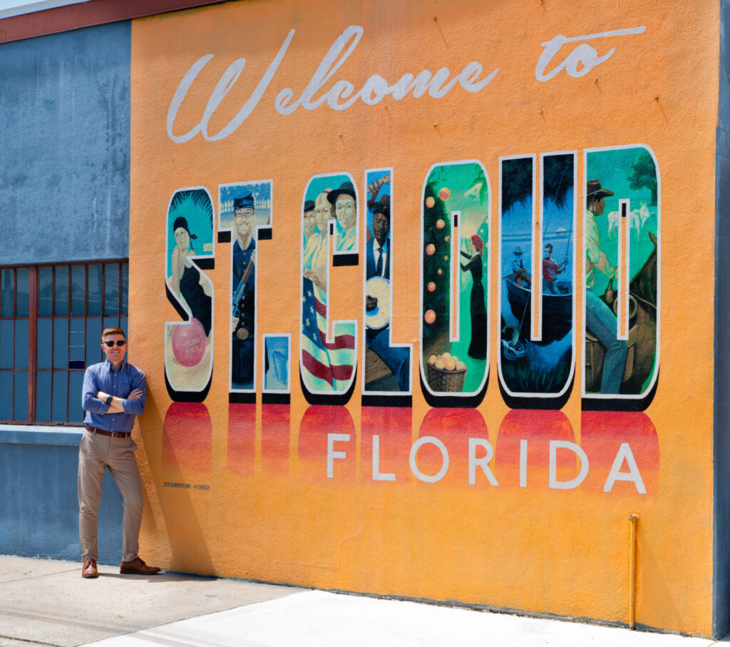 A man in sunglasses stands in front of a mural reading “Welcome to St. Cloud Florida,” with each letter depicting local scenes, on a sunny day—capturing the vibrant spirit that inspires St. Cloud web design and creativity.