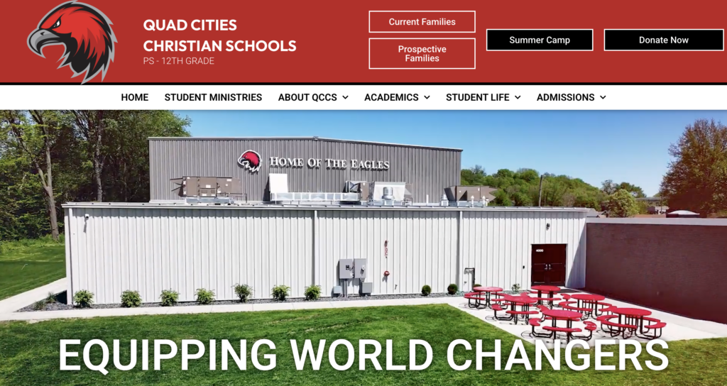 A white metal school building with “Home of the Eagles” written on it, red eagle logo, outdoor red tables, and a green lawn in front. A Quad Cities Christian Schools banner is at the top—an inspiring addition to any Website Design Portfolio highlighting impactful educational environments.