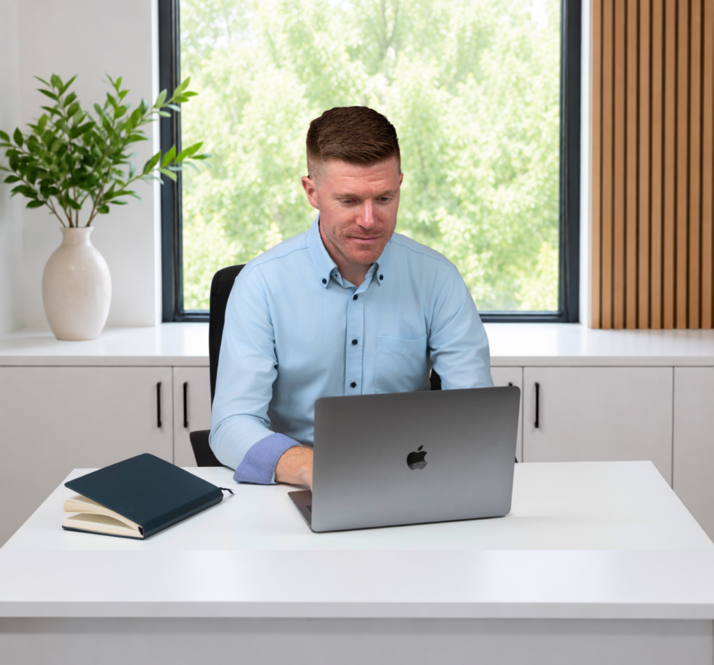 A man in a light blue shirt sits at a white desk, working on his laptop as he prepares for a Free Website Strategy Call. A closed notebook and pen are beside him, ready for taking notes. Behind him, there is a window with greenery outside and a potted plant on a cabinet.