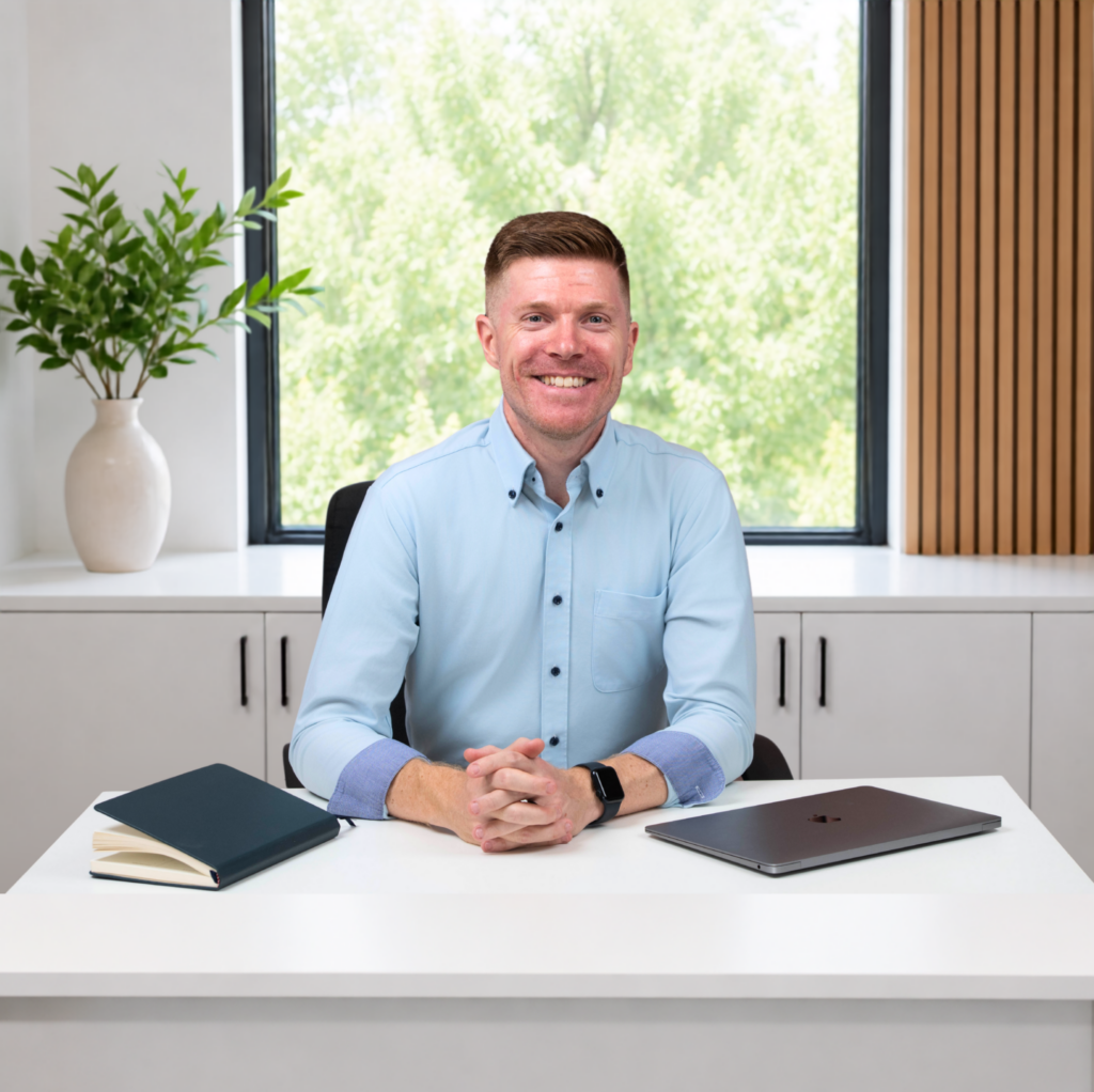 A smiling man in a light blue shirt sits at a white desk with a closed laptop and notebook, working for Anchor Online Web Design in a modern office with a large window and green plant in the background.