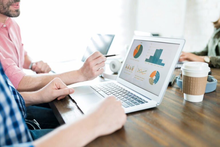 Two people sit at a table, one holding a pen, looking at colorful charts and graphs displayed on a laptop screen as they discuss their mid-year business goals. A disposable coffee cup is nearby.